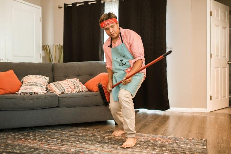 A Man Wearing a Pink Long Sleeves and Blue Apron Holding a Broom While Lying Down on a Couch