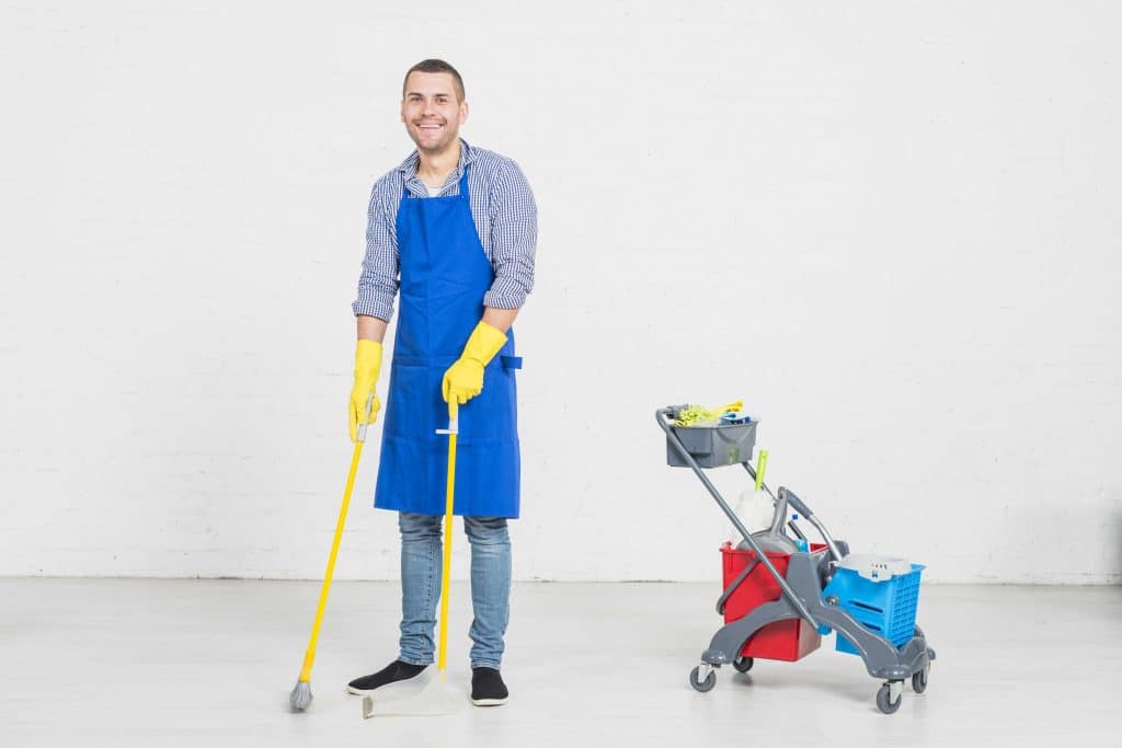 Homme de ménage, balai et pelle en main et souriant. Son chariot contenant son équipement de nettoyage de bureau est à sa gauche.